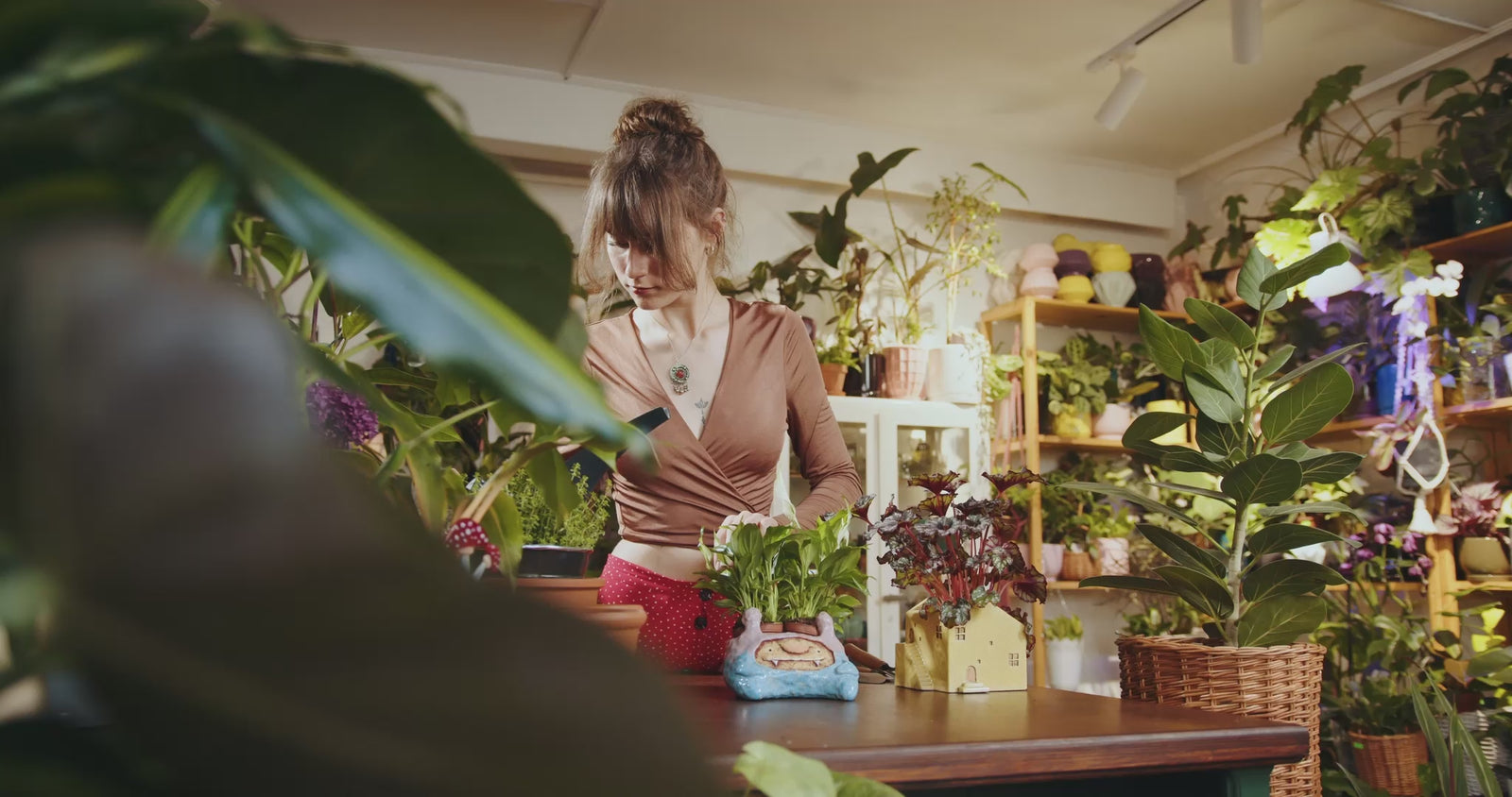 A woman inside a greenhouse filled with plans as she waters one on a table in front of her.